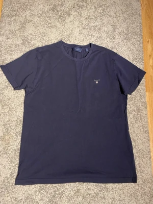 Mörkblå t-shirt från Gant - En stilren mörkblå t-shirt från Gant med liten logotyp på bröstet. Klassisk rund halsringning och korta ärmar. Tillverkad i mjuk bomull som känns skön mot huden. Perfekt för en avslappnad och snygg look.