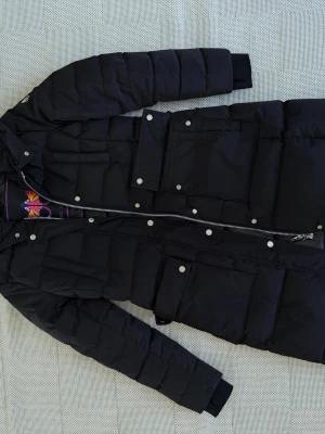 Svart lång dunjacka från Moose knuckels - Säljer en svart lång dunjacka från Moose knuckels med stora fickor, dragkedja och tryckknappar. Jackan har huva och ribbade muddar vid ärmsluten. Perfekt för kalla vinterdagar och riktigt snygg streetstil.