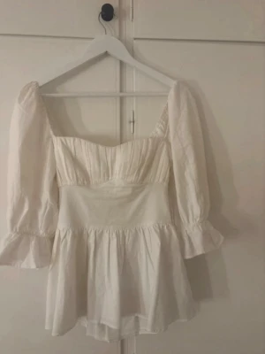 Vit puffärmad blus med rosett - Supersöt vit blus med puffiga långärmar och volangdetaljer. Blusen har en markerad midja och en stor rosett i ryggen som ger en romantisk vibe. Tillverkad i ett lätt, vävt tyg som känns luftigt och skönt. Perfekt att styla med jeans eller kjol.