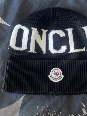 Svart Moncler mössa med logga - Säljer en svart ribbstickad mössa från Moncler med vit text och klassisk Moncler-logga framtill. Mössan har uppvikt kant och är tillverkad i ett mjukt, stretchigt material som sitter skönt på huvudet. Perfekt accessoar för att lyfta din outfit.