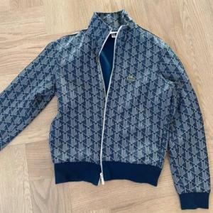 Lacoste - Snygg blå vindjacka från Lacoste med hel dragkedja och hög krage. Jackan har ett mönster med Lacoste-loggor över hela tyget och ribbade muddar i midja och ärmslut. Perfekt för dig som gillar sportig och trendig stil.