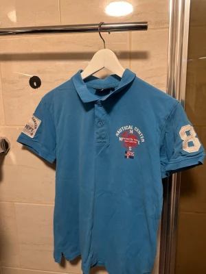 Blå Nautical Center pikétröja - Säljer en blå pikétröja från Nautical Center med tryck och broderade detaljer på bröstet och ärmen. Tröjan har korta ärmar, krage och knappar framtill. Perfekt för en sportig och avslappnad stil.