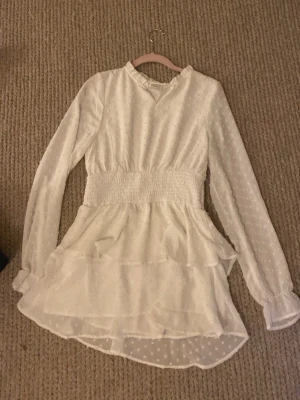 Vit volangklänning med lång ärm - Söt vit klänning med volangdetaljer och smock i midjan. Klänningen har långärmade ärmar med diskreta prickar i tyget och en lätt rynkad halsringning. Perfekt för sommarens alla tillfällen.