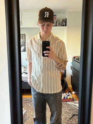 Beige randig skjorta med kort ärm - Säljer en beige skjorta med bruna och vita ränder. Skjortan har korta ärmar, klassisk krage och bröstficka. Perfekt för dig som gillar en avslappnad och stilren look. Materialet känns lätt och bomulligt.