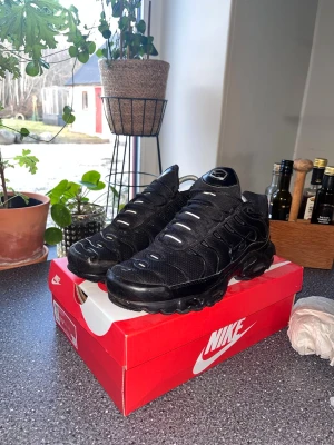 Nike Air Max Plus helsvarta sneakers - Nike Air Max Plus sneakers i helsvart med mesh- och syntetmaterial. Skorna har den ikoniska vågiga designen, synlig Air-dämpning i sulan och reflekterande detaljer. Perfekta för dig som gillar streetwear och vill ha en clean look.