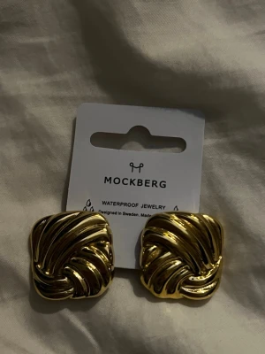 Mockberg Deco guldörhängen - Stilrena örhängen i guldpläterad metall från Mockberg. Fyrkantig form med snirkligt, vågigt mönster som ger en lyxig känsla. Perfekta för dig som gillar statement-smycken och vill sticka ut med en elegant touch.