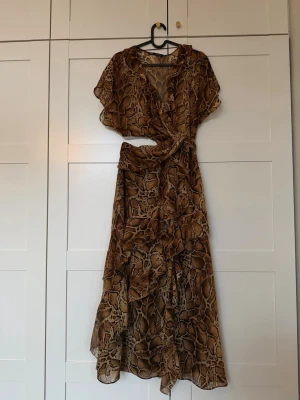 Brun omlottklänning med ormmönster - Säljer en brun långklänning med omlottmodell och volangdetaljer. Klänningen har ett snyggt ormmönster, korta ärmar och v-ringad hals. Materialet är tunt och luftigt, perfekt för varma dagar. Passar dig som vill sticka ut med en trendig look. Använd vid ett tillfälle. Som ny. 