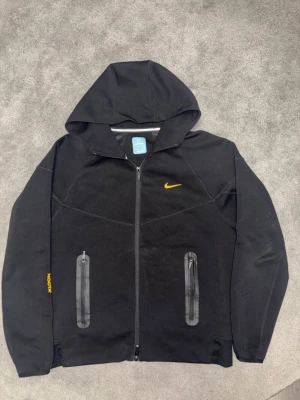 Nike Nocta zipper - Säljer en svart hoodie från Nike med hel dragkedja framtill och två fickor med dragkedja. Hoodien har gul Nike-logga på bröstet och detaljer i gult på ärmen och ryggen. Tillverkad i mjukt material och har en klassisk huva för extra stilpoäng.