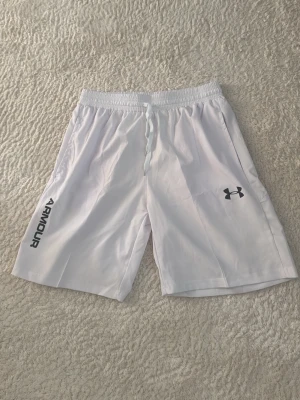 Vita träningsshorts från Under Armour - Säljer ett par vita träningsshorts från Under Armour med elastisk midja och logga på benet. Lätta och luftiga, perfekta för sport eller gym. Klassisk passform och stilren design med tryckt text på sidan. (Aldrig använda)
