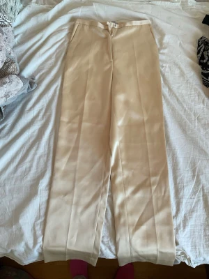 Beiga raka kostymbyxor - Snygga beiga kostymbyxor med hög midja och raka ben. Byxorna har pressveck framtill och stängs med dragkedja och hake. Perfekta för en stilren och clean look.