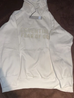 Beige hoodie Essentials Fear of God - Snygg beige hoodie från Essentials Fear of God med stor huva och magficka. Hoodien har tryckt text på bröstet och är gjord i mjukt bomullsmaterial. Perfekt för en avslappnad och trendig stil.