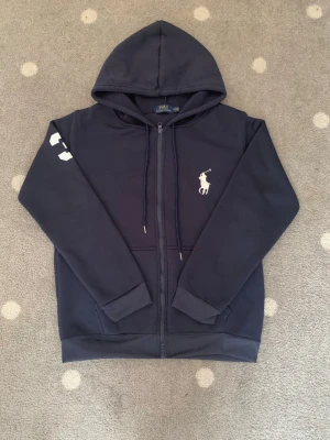 Mörkblå hoodie med dragkedja Polo Ralph Lauren - Mörkblå Polo Ralph Lauren Zip-Up Fleece – Storlek S  Säljer en mörkblå Polo Ralph Lauren zip-up fleece i storlek S. Tröjan är i nyskick, knappt använd och utan några skador eller fläckar.  Den har ett mjukt och skönt fleecematerial på insidan, vilket gör den varm och väldigt bekväm att bära. Tröjan har en större Polo Ralph Lauren-logga på bröstet samt den klassiska siffran 3 på högra ärmen.  Skick: Nyskick / knappt använd Storlek: S Pris: 800kr 