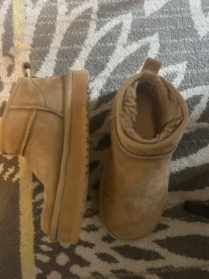 UGG classic ultra mini - Mysiga UGGs i beige mocka med mjukt foder och grov sula. Modellen är låg och har en rund tå samt dragflik baktill. Perfekta för kalla dagar när du vill ha både stil och komfort.
