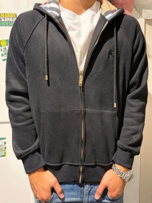 Svart burberry zip  - Svart hoodie med dragkedja framtill, huva med snören och randigt innerfoder. Modellen har två stora fickor framtill och ribbade muddar vid ärmslut och nederkant. Perfekt för en avslappnad och stilren look.