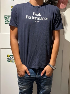 Mörkblå Peak Performance t-shirt - Snygg mörkblå t-shirt från Peak Performance med vit logotyptryck på bröstet. Klassisk rund halsringning och korta ärmar. Perfekt för en avslappnad och stilren look.
