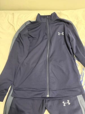 Blått träningsset från Under Armour - Säljer ett blått träningsset från Under Armour i storlek M. Setet består av en zip-jacka och matchande byxor, båda med logga på bröst och ben. Materialet är mjukt och stretchigt, perfekt för träning eller chill. Jackan har hög krage och dragkedja framtill.