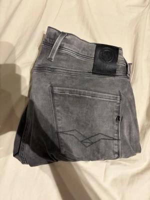 Replay anbass jeans  - Säljer ett par grå tvättade jeans med raka ben och klassisk femficksdesign. Jeansen har en svart patch bak i midjan och diskreta sömmar på bakfickorna. Materialet är denim i bomull och de har en avslappnad look som passar till det mesta.