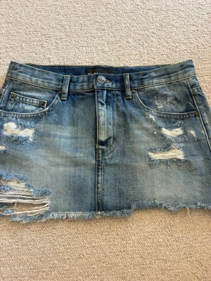 Blå jeanskjol från Ziggy Denim - Säljer en kort jeanskjol från Ziggy Denim i blå tvätt med slitna detaljer och fransig nederkant. Kjolen har klassiska fem fickor, bälteshällor och dragkedja framtill. Perfekt för dig som gillar en avslappnad och trendig stil.💓