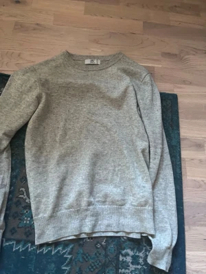 Ljusgrå stickad tröja från BVALDI  - En stilren ljusgrå stickad tröja från BVALDI med rund halsringning och ribbade muddar vid ärmslut och nederkant. Perfekt basplagg i mjukt material som passar till det mesta. Enkel och klassisk design utan mönster. Litet hål