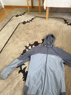 Windbreaker grå från Under Armour - Snygg hoodie från Under Armour i två nyanser av grått med dragkedja framtill och stor huva. Logga på bröstet och stor text på ryggen. Tillverkad i ett mjukt och sportigt material, perfekt för chill eller träning. Elastiska muddar vid ärmsluten och praktiska fickor.