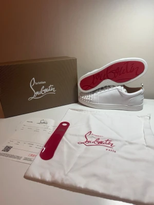 Vita Christian Louboutin sneakers - Snygga vita sneakers från Christian Louboutin med klassisk röd sula och coola nitar på tån. Skorna är låga och har en stilren design i läder. Perfekta för dig som vill sticka ut med exklusiva detaljer och premiumkänsla.