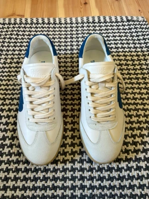 The Gallerist Skor Strl 41 Helt Oanvända  - Snygga vita sneakers från The Gallerist med blå klöverdetalj på sidan och blå häl. Skorna har klassisk låg profil, vita snören och ovandel i mocka och läder. Perfekta för dig som vill ha en clean och stilren look. Låta ingår.
