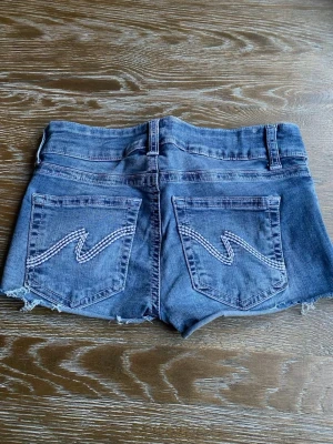 Blå jeansshorts med rå kant - Snygga blå jeansshorts med rå, fransig kant och klassiska fem fickor. Baksidan har coola broderade detaljer på fickorna. Perfekta för varma dagar och enkel att matcha med allt. Midja med dubbla knappar och bälteshällor. jättefina Stockholm stil jeans shorts från Nelly