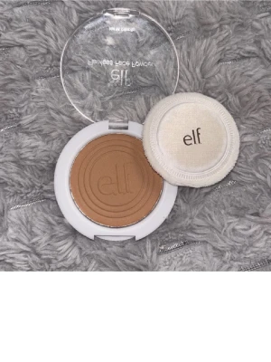 Bronzer från e.l.f. med applikator - Snygg bronzer från e.l.f. i en rund vit dosa med transparent lock. Pudret har en varm, mellanbrun ton och kommer med en mjuk applikator. Perfekt för att ge ansiktet en solkysst look och definiera ansiktsdragen.