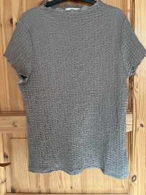 Grå ribbad topp med kort ärm - En enkel grå topp med korta ärmar och ribbad struktur. Materialet är mjukt och stretchigt, perfekt för en avslappnad look. Toppen har diskreta vågiga kanter vid ärmslut och halsringning för en extra detalj.