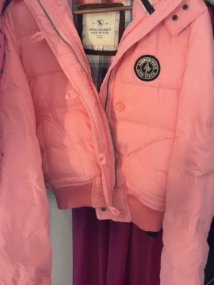 Rosa pufferjacka från Abercrombie - Säljer en rosa pufferjacka från Abercrombie & Fitch med ribbade muddar, dragkedja och knappstängning. Jackan har en patch med logga på bröstet och rutigt innerfoder. Perfekt för kalla vinterdagar.