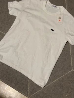 Vit t-shirt från Lacoste - Klassisk vit t-shirt från Lacoste med broderad grön krokodil på bröstet. T-shirten har rund halsringning och korta ärmar. Tillverkad i mjuk bomull och har en regular fit som passar till det mesta.