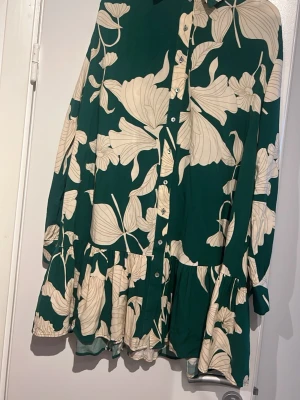 Grön blommig kortklänning med volang - Grön kortklänning med stora beige blommönster, knappar framtill och volang nedtill. Klänningen har lång ärm med knapp vid ärmslut och en avslappnad passform. Perfekt för dig som gillar färg och mönster.