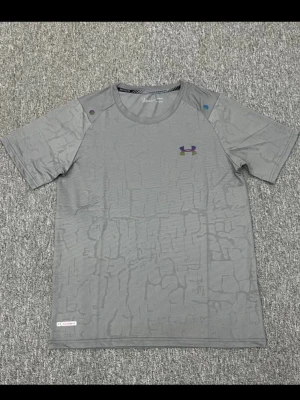 Under Armour träningsset grå/blå - Snyggt träningsset från Under Armour med en grå t-shirt i tekniskt material och diskret mönster samt blå träningsshorts med dragsko och elastisk midja. Båda plaggen har logga på bröst och ben. Perfekt för gym eller löpning.