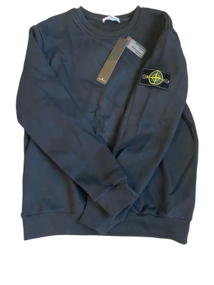 Svart sweatshirt från Stone Island - Snygg svart sweatshirt från Stone Island med klassisk rund hals och ikonisk patch på ärmen. Tröjan har ribbade muddar vid ärmslut och nederkant. Perfekt för dig som gillar streetwear och vill ha en stilren look.