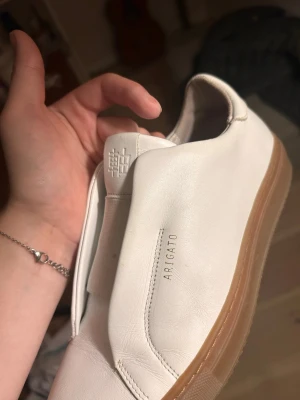 Vita sneakers från Axel Arigato - Stilrena vita slip-on sneakers från Axel Arigato med minimalistisk design. Skorna har ovandel i slätt läder och en tjock, ljusbrun gummisula. Diskret logga på sidan och plös. Perfekta för dig som gillar enkel och clean stil. Väldigt sparsamt använda då de är för små. Är öppen för att diskutera pris💕