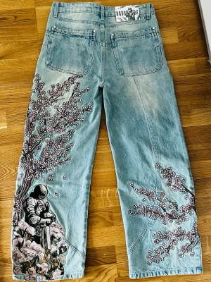 Baggy ljusblå jeans med tryck - Unika ljusblå jeans med bred passform och coola tryck av blommor och en figur på benen och baksidan. Spillrans nya knappt använd 