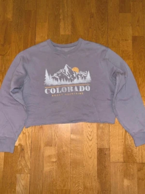Lila cropped Colorado  - Cropped  i lila med tryck av berg, sol och texten 'Colorado Rocky Mountains' på bröstet. Tröjan har långa ärmar, ribbade muddar och rund halsringning. Perfekt för en avslappnad och trendig look.