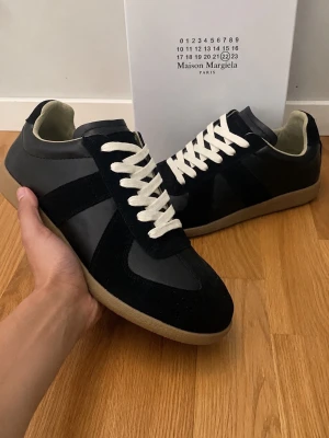 Maison Margiela svarta - Maison Margiela sneakers i svart läder och mocka med vita skosnören och beige gummisula. Klassisk low-top siluett med stilrena detaljer och kontrasterande materialval. Perfekta för dig som gillar minimalistisk och tidlös design.