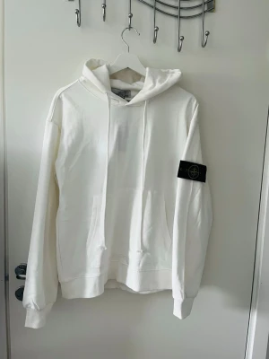 Vit hoodie med patch på ärmen - Snygg vit hoodie med huva och känguruficka framtill. Har en svart patch med logga på vänster ärm som ger en cool detalj. Mjuk och skön bomullskänsla, perfekt för en avslappnad streetwear-look.