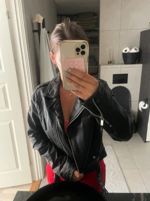 Svart klassisk skinnjacka - Snygg svart skinnjacka med dragkedja och klassisk bikerkrage. Jackan har detaljerade sömmar och fickor med dragkedja, vilket ger en cool och tidlös look. Perfekt att slänga över en hoodie eller t-shirt för en edgy stil.