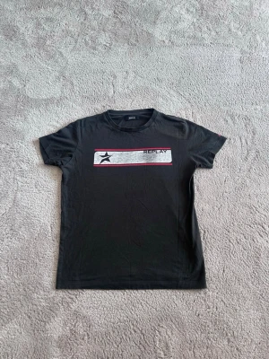 Svart t-shirt från Replay - Svart t-shirt från Replay med korta ärmar och rund hals. På bröstet finns en vit och röd rektangel med en svart stjärna och Replay-loggan. Perfekt för en avslappnad och stilren look.