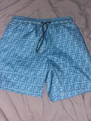 Blå mönstrade Fendi shorts med snörning - Säljer ett par ljusblå Fendi  shorts med all-over logomönster och svart snörning i midjan. Shortsen har elastisk midja och sidofickor, perfekta för sommaren. Materialet känns lätt och bekvämt, passar till chill dagar eller stranden.