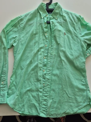 Mintgrön skjorta med knappar(Ralph Lauren) - Snygg mintgrön skjorta med klassisk krage och knappar framtill. Skjortan har lång ärm och en liten broderad logga på bröstet. Tillverkad i mjuk bomull som känns skön mot huden. Perfekt för dig som gillar stilrena plagg med en fräsch färg. En liten fläck fram som knappt syns(kolla bild)