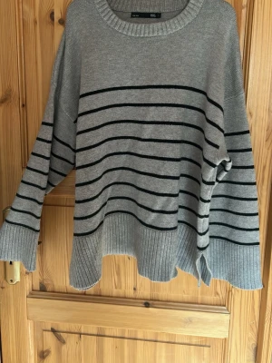 Grå oversize stickad tröja med ränder - Säljer en grå stickad tröja i oversize-modell med svarta horisontella ränder. Tröjan har rund halsringning, långa ärmar och bred ribbstickad kant nedtill samt vid ärmsluten. Perfekt för en avslappnad och trendig look.