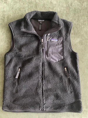 Svart fleeceväst från Patagonia - Mysig svart fleeceväst från Patagonia med hög krage och dragkedja framtill. Västen har två sidofickor med dragkedja och en bröstficka i nylon med logga. Perfekt för lager-på-lager och outdoor-vibe.