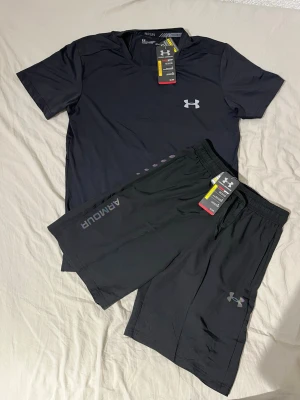 Svart träningsset från Under Armour - Snyggt svart träningsset från Under Armour med t-shirt och shorts. T-shirten har en diskret logga på bröstet och shortsen har elastisk midja, dragsko och logga på benet. Pris kan diskuteras vid snabbaffär!🤗