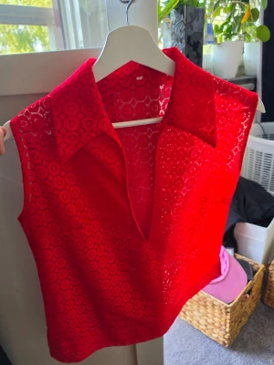 Röd spetsig ärmlös linnetopp - Snygg röd ärmlös linnetopp med spetsdetaljer och krage. Toppen har v-ringning och är gjord i ett genombrutet spetsmaterial som ger en cool och trendig look. Perfekt att styla med jeans eller kjol för en extra touch.