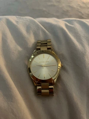 Guldig klocka från Michael Kors - Stilren klocka från Michael Kors i guldigt metallarmband och rund urtavla. Urtavlan är ljus med minimalistiska markeringar och utan siffror. Klassisk design som passar till många olika stilar.