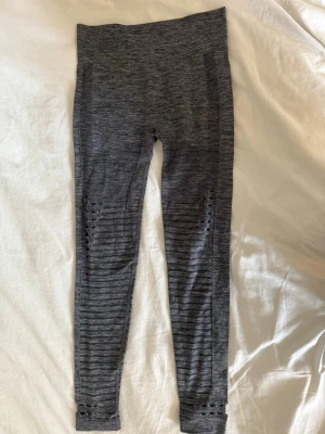 Gråa träningsbyxor  -  Grå träningsbyxor med sömlös design och diskreta håldetaljer längs benen. Hög midja och stretchigt material som sitter skönt på kroppen. Storlek XS/S. Slitning längst ner i benet. 