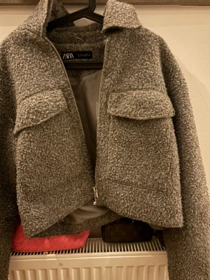 Grå teddyjacka från ZARA - Säljer en grå teddyjacka från ZARA med krage, dragkedja och två stora bröstfickor med lock. Jackan har en mjuk, fluffig yta och är fodrad på insidan. Perfekt för dig som vill ha en trendig och mysig jacka till hösten eller vintern. Känns som Xs/S